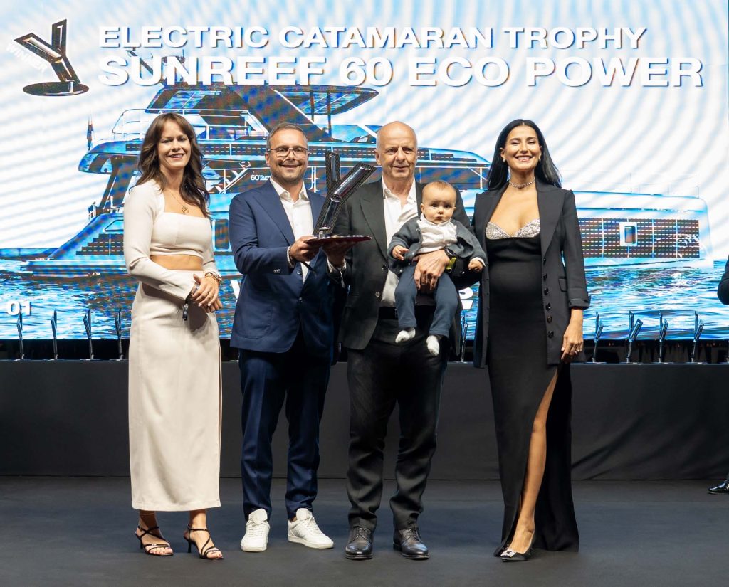 Group of people posing for a photo at the Sunreef Yachts Electric Catamaran Trophy event, with one person holding a trophy and another holding a baby.