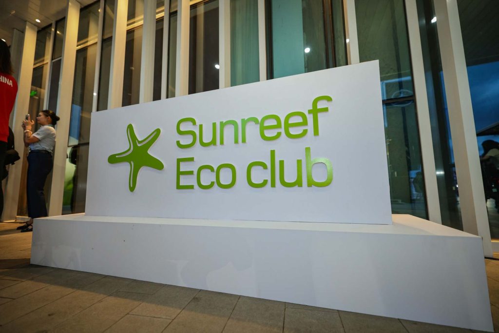 The image depicts a signage board for 'Sunreef Eco Club' with the Sunreef Yachts logo, placed in front of a building with glass facade. A person is seen standing in the background to the left.