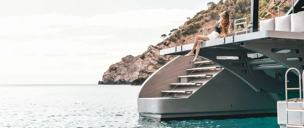 A woman sitting on the steps of a Sunreef Yacht, with the ocean and a rocky hill in the background.