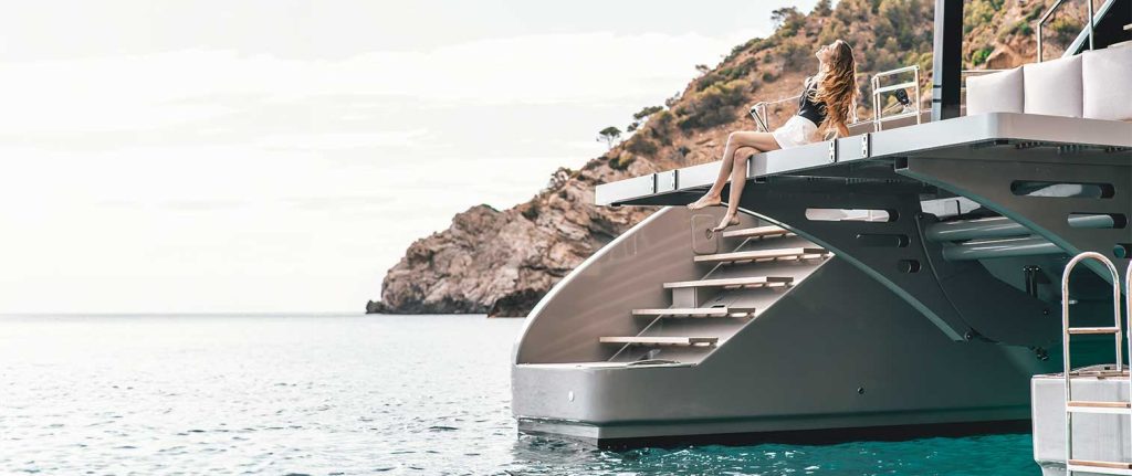 A woman sitting on the steps of a Sunreef Yacht, with the ocean and a rocky hill in the background.